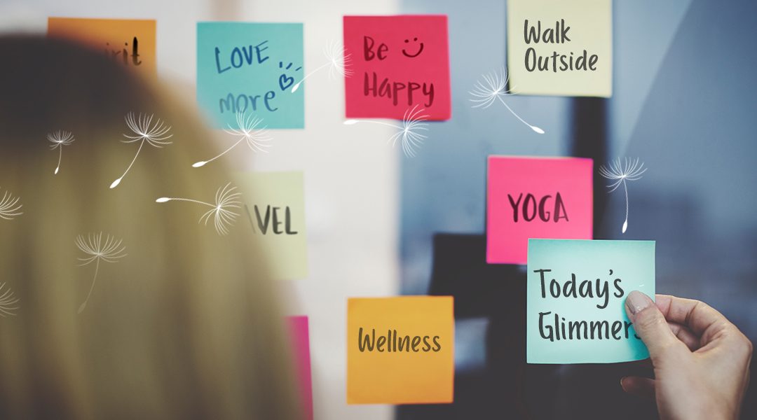 A woman placing mindful notes on a board: Love more, Be happy, go outside, Yoga, wellness, and Today's Glimmers.
