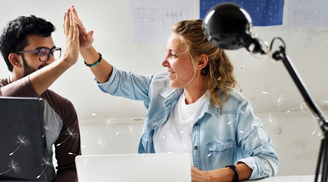 Two colleagues sit at a desk, smiling and giving each other a high-five across their laptops. One is a man with glasses and a beard, and the other is a woman with a ponytail wearing a denim jacket. Dandelion seed illustrations float across the image.