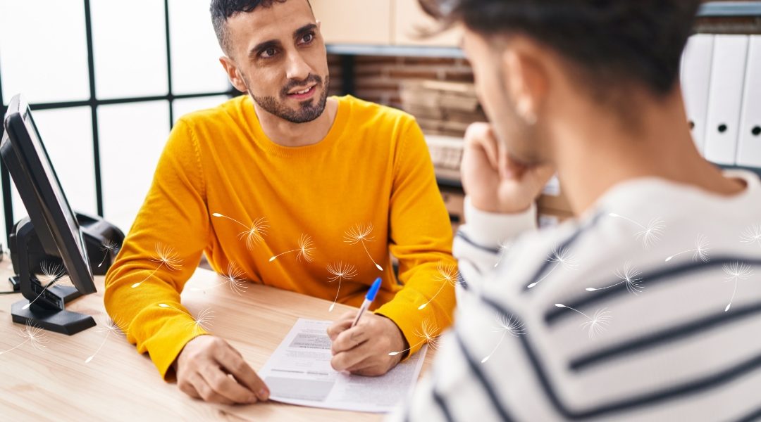 Two people sit across from each other at a desk in a well-lit office, engaged in a conversation. One person in a yellow sweater is holding a pen and a paper, while the other, in a striped shirt, listens thoughtfully. Dandelion seeds float across the image, symbolizing the Specialisterne logo.