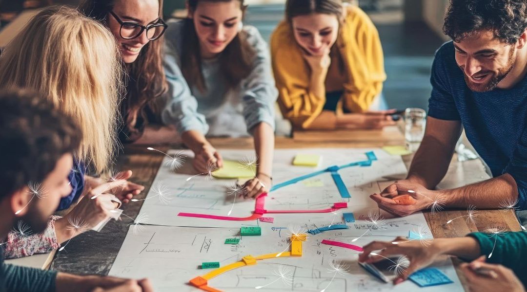 A group of six people collaborate around a large table covered with paper plans, sticky notes, and colorful markers. They are smiling and leaning in, actively engaged in brainstorming. Light dandelion graphics float across the image, symbolizing inclusion and ideas spreading.